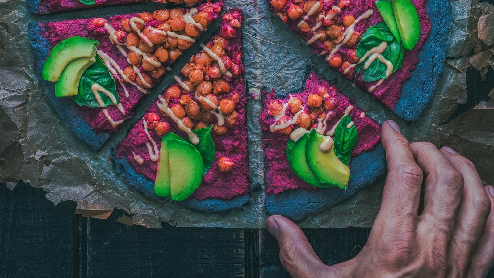 Topview auf eine ungewöhnliche Pizza mit lila Boden, belegt mit Kichererbsen und Avocado.