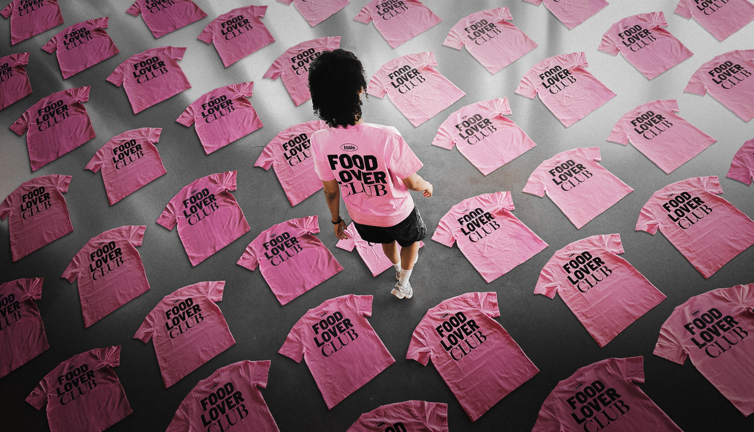 Topview auf viele rosafarbene T-Shirts, die auf einem Betonboden liegen; eine junge Frau, die das gleiche T-Shirt trägt läuft zwischen den Hemden durchs Bild.