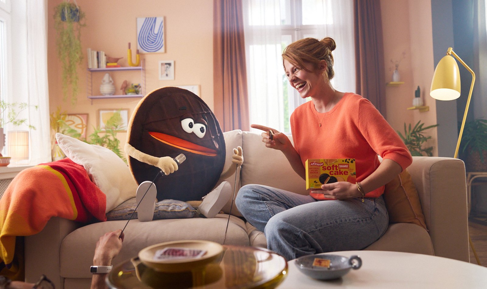 Eine überdimensional große Soft Cake Puppe sitzt lachend mit einer jungen Frau auf einer Couch.