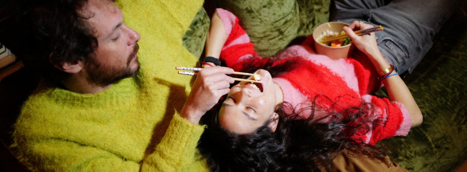 Ein junges Paar liegt auf der Couch, er füttert sie mit Stäbchen.