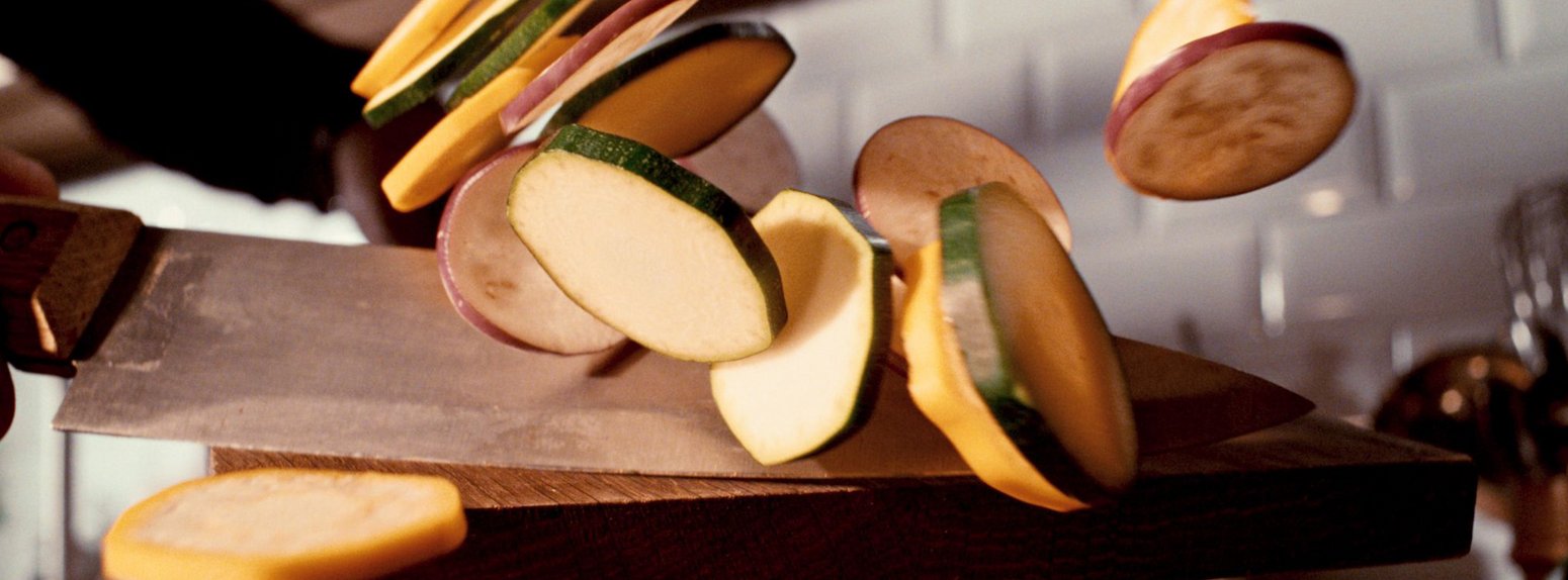 Closeup auf geschnittene Zucchini, die mit einem Kochmesser vom Brett in die Pfanne geschoben werden.