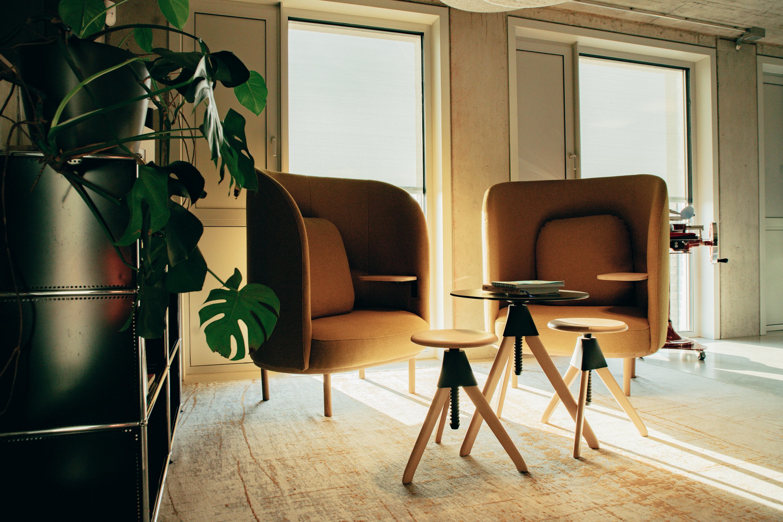 Hell erleuchtete Leseecke in einem modernen Büro mit zwei gelben Sesseln, Holzhockern und einer großen Monstera-Pflanze.