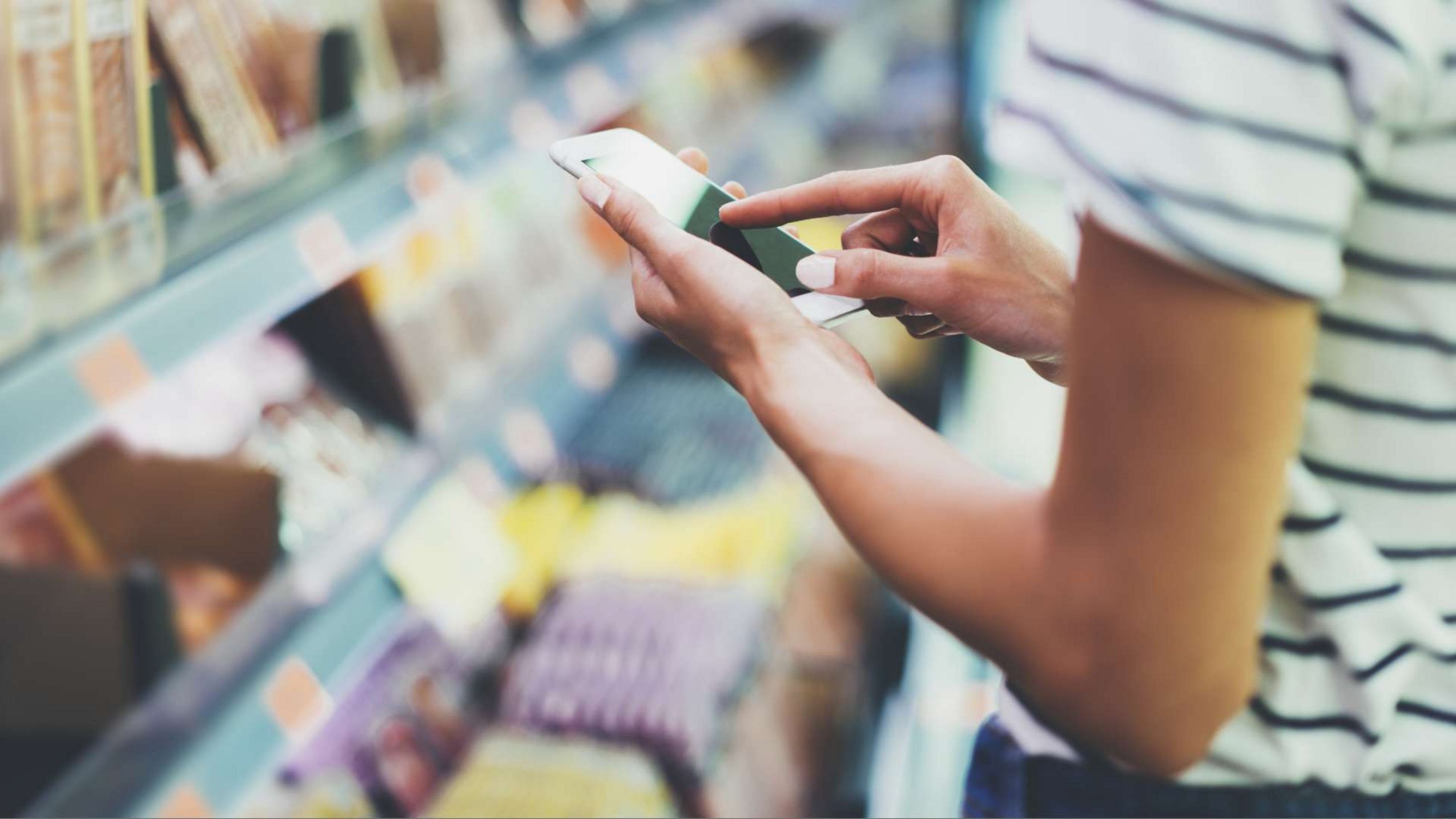 Closeup auf die Hände einer Frau, die vor einem Supermarktregal etwas im Smartphone checkt.