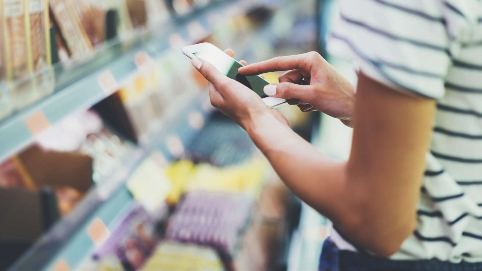 Closeup auf die Hände einer Frau, die vor einem Supermarktregal etwas im Smartphone checkt.