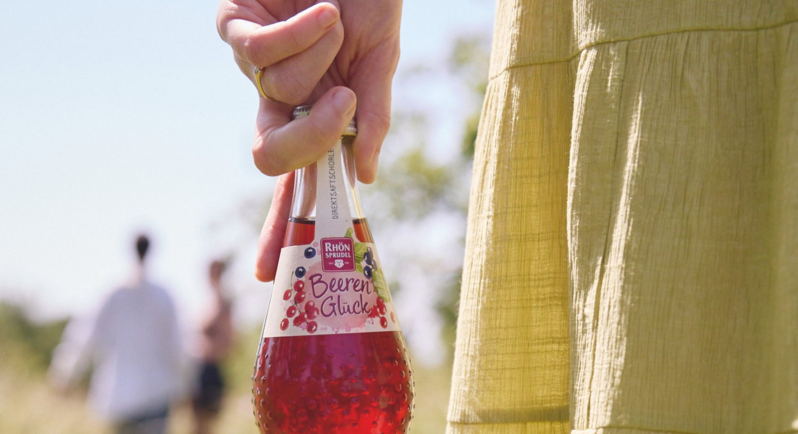 Closeup in sommerlichem Setting auf eine Flasche Beerenglück, die von einer Frau locker am Flaschenhals gehalten wird.