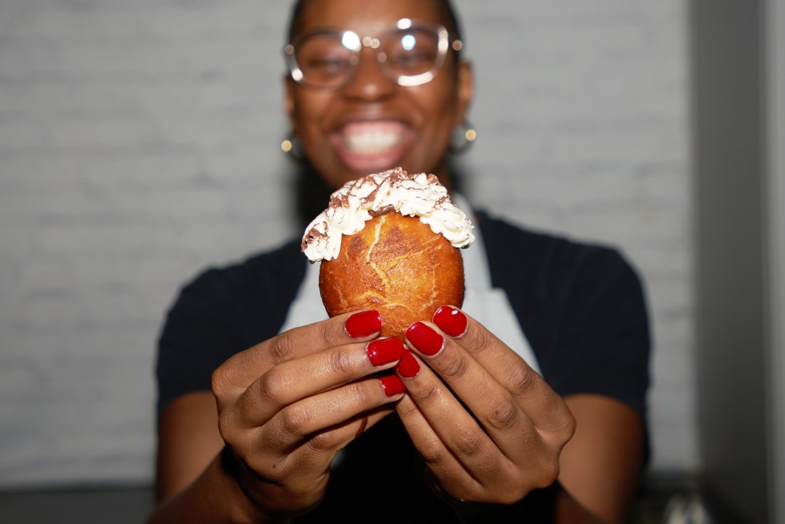 Eine Kollegin hält lächelnd ein Gebäck in die Kamera, nur eine von vieleln leckeren Aktionen des Feelgood-Teams.