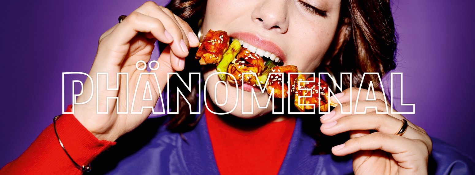 Closeup einer jungen Frau, die genüsslich in einen asiatischen Hähnchenspieß beißt, darüber das Wort "Phänomenal".